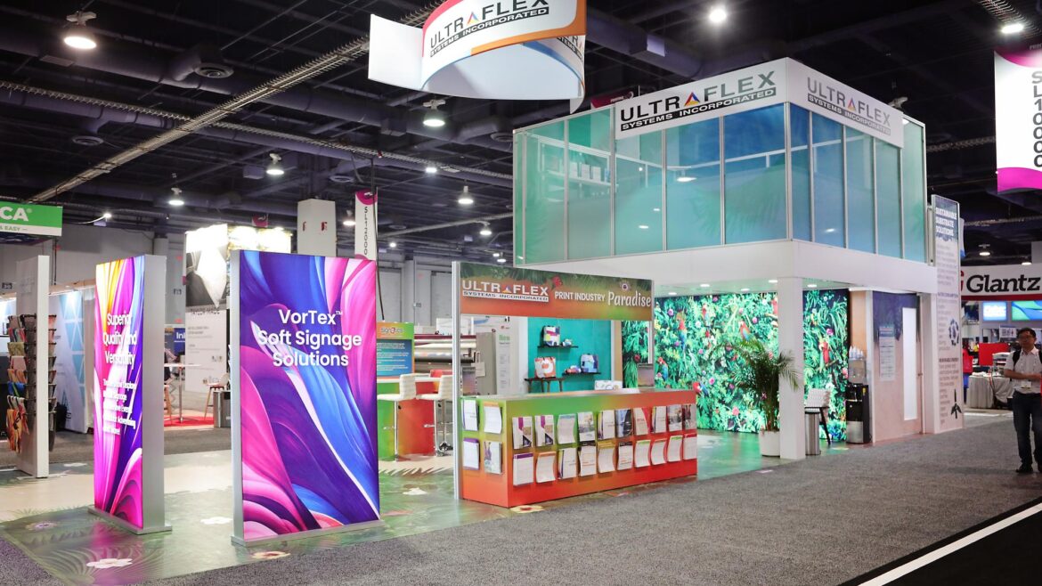 UltraFlex Systems trade show booth at PRINTING United, with backlit and front-lit VorTex soft signage SEG panels in aluminum frames